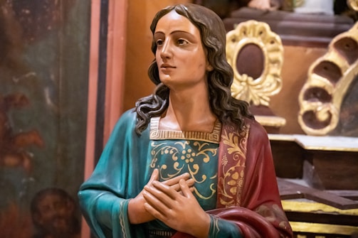 A detailed religious statue of a person with long hair, wearing a richly decorated robe with vibrant colors. The figure has a serene and contemplative expression, and the background features ornate, golden decorative elements.