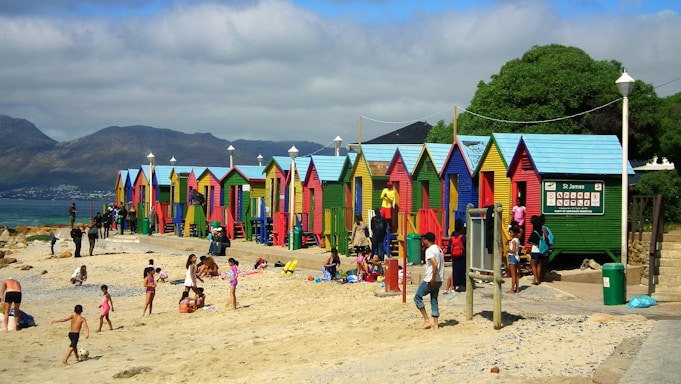Brightly colored beach huts line the edge of a sandy beach, while several people enjoy leisure activities such as sunbathing and playing. The beach appears lively with numerous families and individuals interacting. In the background, mountains rise under a partly cloudy sky, creating a picturesque scene.