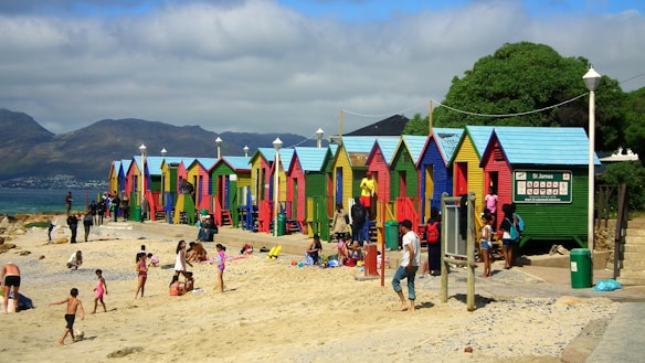 Brightly colored beach huts line the edge of a sandy beach, while several people enjoy leisure activities such as sunbathing and playing. The beach appears lively with numerous families and individuals interacting. In the background, mountains rise under a partly cloudy sky, creating a picturesque scene.
