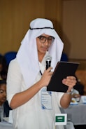 A person wearing traditional attire, including a white headscarf and glasses, is holding a microphone and reading from a tablet or notebook. They are standing in a conference-like setting with a name tag and a small Saudi Arabian flag visible on the table nearby.