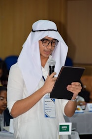 A person wearing traditional attire, including a white headscarf and glasses, is holding a microphone and reading from a tablet or notebook. They are standing in a conference-like setting with a name tag and a small Saudi Arabian flag visible on the table nearby.