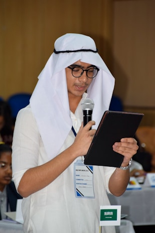 A person wearing traditional attire, including a white headscarf and glasses, is holding a microphone and reading from a tablet or notebook. They are standing in a conference-like setting with a name tag and a small Saudi Arabian flag visible on the table nearby.