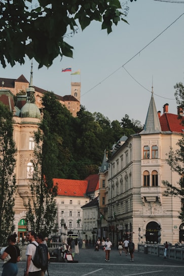 A bustling town square with historic European architecture, including ornate buildings with red and gray rooftops. A castle on a hill is visible in the background, surrounded by lush greenery. People are walking and socializing in the stone-paved area below.