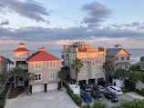 A group of three modern beachfront houses with red and gray roofs are situated near the ocean under a partly cloudy sky. The sunlight casts a warm glow on the buildings. Several cars are parked in front, and there are palm trees in the vicinity.