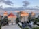 A group of three modern beachfront houses with red and gray roofs are situated near the ocean under a partly cloudy sky. The sunlight casts a warm glow on the buildings. Several cars are parked in front, and there are palm trees in the vicinity.