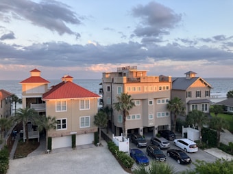 A group of three modern beachfront houses with red and gray roofs are situated near the ocean under a partly cloudy sky. The sunlight casts a warm glow on the buildings. Several cars are parked in front, and there are palm trees in the vicinity.