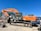 An orange excavator with a large arm and bucket is parked on a dirt surface. The machine is branded with 'Hitachi' and has visible tracks suitable for maneuvering on rugged terrains. In the background, there's a corrugated metal structure and some trees against a clear blue sky.