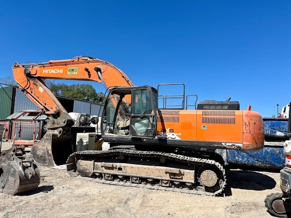 An orange excavator with a large arm and bucket is parked on a dirt surface. The machine is branded with 'Hitachi' and has visible tracks suitable for maneuvering on rugged terrains. In the background, there's a corrugated metal structure and some trees against a clear blue sky.