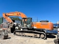 An orange excavator with a large arm and bucket is parked on a dirt surface. The machine is branded with 'Hitachi' and has visible tracks suitable for maneuvering on rugged terrains. In the background, there's a corrugated metal structure and some trees against a clear blue sky.