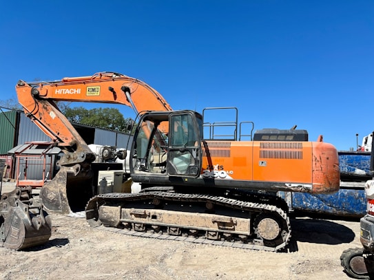 An orange excavator with a large arm and bucket is parked on a dirt surface. The machine is branded with 'Hitachi' and has visible tracks suitable for maneuvering on rugged terrains. In the background, there's a corrugated metal structure and some trees against a clear blue sky.