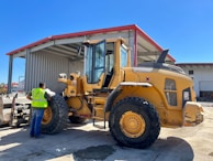 A construction worker wearing a safety vest stands beside a large yellow front-end loader in an industrial setting. The loader is parked near a large metal warehouse with a red roof, and the man appears to be inspecting it. The scene is well-lit, suggesting it is a sunny day.