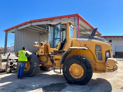 A construction worker wearing a safety vest stands beside a large yellow front-end loader in an industrial setting. The loader is parked near a large metal warehouse with a red roof, and the man appears to be inspecting it. The scene is well-lit, suggesting it is a sunny day.