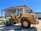 A construction worker wearing a safety vest stands beside a large yellow front-end loader in an industrial setting. The loader is parked near a large metal warehouse with a red roof, and the man appears to be inspecting it. The scene is well-lit, suggesting it is a sunny day.