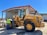 A construction worker wearing a safety vest stands beside a large yellow front-end loader in an industrial setting. The loader is parked near a large metal warehouse with a red roof, and the man appears to be inspecting it. The scene is well-lit, suggesting it is a sunny day.