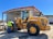 A construction worker wearing a safety vest stands beside a large yellow front-end loader in an industrial setting. The loader is parked near a large metal warehouse with a red roof, and the man appears to be inspecting it. The scene is well-lit, suggesting it is a sunny day.
