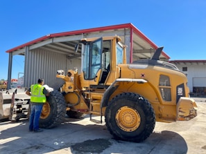 A construction worker wearing a safety vest stands beside a large yellow front-end loader in an industrial setting. The loader is parked near a large metal warehouse with a red roof, and the man appears to be inspecting it. The scene is well-lit, suggesting it is a sunny day.