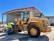 A construction worker wearing a safety vest stands beside a large yellow front-end loader in an industrial setting. The loader is parked near a large metal warehouse with a red roof, and the man appears to be inspecting it. The scene is well-lit, suggesting it is a sunny day.