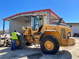 A construction worker wearing a safety vest stands beside a large yellow front-end loader in an industrial setting. The loader is parked near a large metal warehouse with a red roof, and the man appears to be inspecting it. The scene is well-lit, suggesting it is a sunny day.