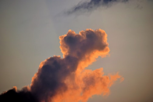 A cloud illuminated by the soft, warm glow of sunrise or sunset, with vibrant orange and pink edges against a calm, muted sky. The cloud shapes appear fluffy and intricate, resembling a natural sculpture in the sky.