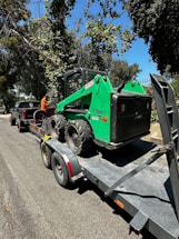 A green Bobcat skid steer loader is secured on a flatbed trailer parked on a road. The trailer is attached to a black pickup truck. The scene is surrounded by trees and greenery, and the sky is clear and blue.