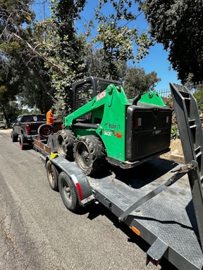 A green Bobcat skid steer loader is secured on a flatbed trailer parked on a road. The trailer is attached to a black pickup truck. The scene is surrounded by trees and greenery, and the sky is clear and blue.