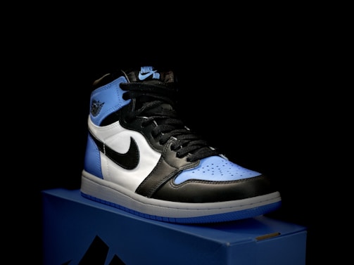 A stylish athletic shoe is placed on a shoebox, featuring a combination of black, white, and light blue. The shoe design includes a prominent logo on the side and a high-top silhouette.