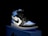 A stylish athletic shoe is placed on a shoebox, featuring a combination of black, white, and light blue. The shoe design includes a prominent logo on the side and a high-top silhouette.