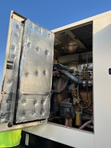 An industrial engine is partially visible within a large enclosure. The open panel is lined with silver insulation material, featuring a quilted pattern. Various mechanical components such as pipes, tubes, and a fan are evident. A small section of a person wearing a yellow shirt is present at the bottom left corner.