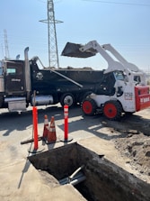 A construction site featuring a white Bobcat loader with red wheels unloading dirt into a black dump truck. In the foreground, there is a trench with visible pipes and several orange safety cones and bollards indicating a work zone. In the background, electrical power lines and a pylon are visible against a clear blue sky.