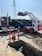 A construction site featuring a white Bobcat loader with red wheels unloading dirt into a black dump truck. In the foreground, there is a trench with visible pipes and several orange safety cones and bollards indicating a work zone. In the background, electrical power lines and a pylon are visible against a clear blue sky.