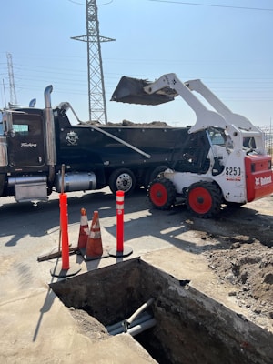 A construction site featuring a white Bobcat loader with red wheels unloading dirt into a black dump truck. In the foreground, there is a trench with visible pipes and several orange safety cones and bollards indicating a work zone. In the background, electrical power lines and a pylon are visible against a clear blue sky.
