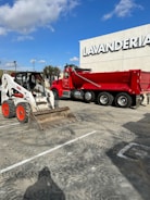 A red dump truck and a white skid steer loader are parked in a lot in front of a building with the word 'Lavanderia' displayed prominently. The sky above is clear with some clouds, and there is a streetlamp and some trees in the background.