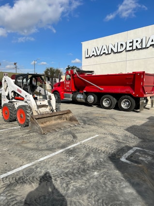 A red dump truck and a white skid steer loader are parked in a lot in front of a building with the word 'Lavanderia' displayed prominently. The sky above is clear with some clouds, and there is a streetlamp and some trees in the background.