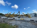 A construction yard with multiple yellow telescopic handlers arranged in a row. They are parked on an open area with a pile of gravel in the center. A wire fence runs across the foreground, and there's a building with an open door in the background under a clear blue sky with scattered clouds.