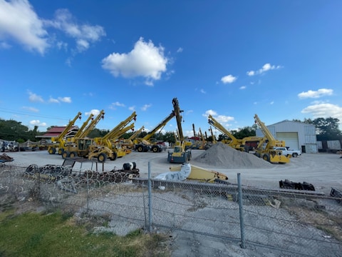 A construction yard with multiple yellow telescopic handlers arranged in a row. They are parked on an open area with a pile of gravel in the center. A wire fence runs across the foreground, and there's a building with an open door in the background under a clear blue sky with scattered clouds.