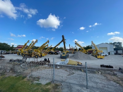 A construction yard with multiple yellow telescopic handlers arranged in a row. They are parked on an open area with a pile of gravel in the center. A wire fence runs across the foreground, and there's a building with an open door in the background under a clear blue sky with scattered clouds.