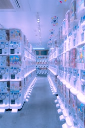 A brightly lit aisle lined with numerous vending machines on both sides, displaying various colorful items inside them. The machines have a modern, clean design, and the floor is carpeted. The ceiling has several spotlights and decorations with vibrant patterns are hanging overhead.