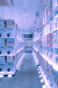 A brightly lit aisle lined with numerous vending machines on both sides, displaying various colorful items inside them. The machines have a modern, clean design, and the floor is carpeted. The ceiling has several spotlights and decorations with vibrant patterns are hanging overhead.