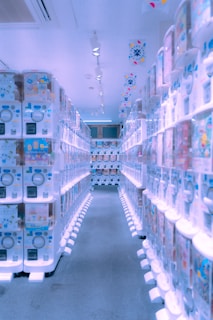 A brightly lit aisle lined with numerous vending machines on both sides, displaying various colorful items inside them. The machines have a modern, clean design, and the floor is carpeted. The ceiling has several spotlights and decorations with vibrant patterns are hanging overhead.