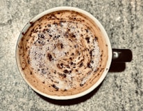 A cup of coffee with frothy milk on the surface, topped with chocolate powder or cocoa. The pattern on top is swirled and speckled with the powder, creating a textured appearance. The cup is placed on a speckled countertop.