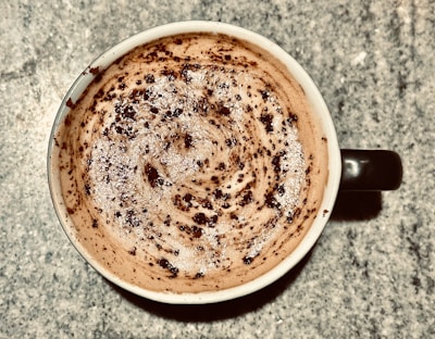 A cup of coffee with frothy milk on the surface, topped with chocolate powder or cocoa. The pattern on top is swirled and speckled with the powder, creating a textured appearance. The cup is placed on a speckled countertop.