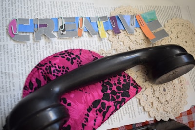 A cut-out collage with colorful letters forming the word 'CREATIVE' is placed on top of a page of text. A black telephone handset rests on a vibrant, pink fabric featuring a black, leopard-like print. Underneath are lace doilies.