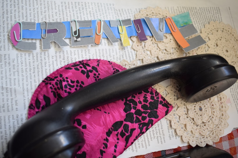 A cut-out collage with colorful letters forming the word 'CREATIVE' is placed on top of a page of text. A black telephone handset rests on a vibrant, pink fabric featuring a black, leopard-like print. Underneath are lace doilies.