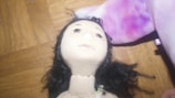A close-up view of a fabric doll featuring a detailed face with black hair. The doll is placed on a wooden surface, and there is a purple and pink plush item partially visible beside it.
