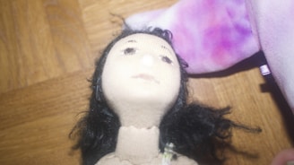 A close-up view of a fabric doll featuring a detailed face with black hair. The doll is placed on a wooden surface, and there is a purple and pink plush item partially visible beside it.