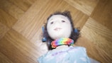 A handmade cloth doll with a soft face and curly dark hair, adorned with a colorful rainbow bracelet around its neck. It wears a patterned dress in pastel shades against a wooden parquet floor background.