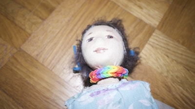 A handmade cloth doll with a soft face and curly dark hair, adorned with a colorful rainbow bracelet around its neck. It wears a patterned dress in pastel shades against a wooden parquet floor background.