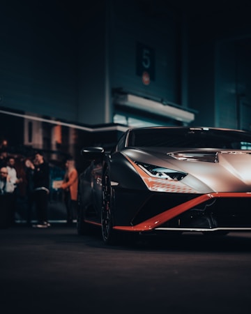 A sleek, modern sports car displayed in a dimly lit setting, with a matte finish and striking design features. The background shows a few people, slightly out of focus, suggesting an indoor automotive event or showroom atmosphere.