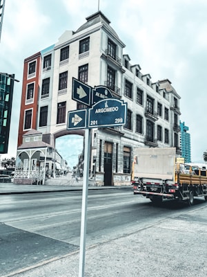 A multi-story building with classic architectural design stands at the corner of a street. A street sign in the foreground indicates directions for Argomedo Avenue. There is a mural on the building's side depicting a historical street scene. A yellow delivery truck is driving by on the road, while other modern buildings are visible in the background.