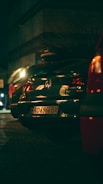 A nighttime scene on a street with several parked cars. A Volkswagen with a visible license plate is at the center, illuminated by streetlights, while other vehicles are partially visible. The surrounding environment is dimly lit, contributing to a moody ambiance.